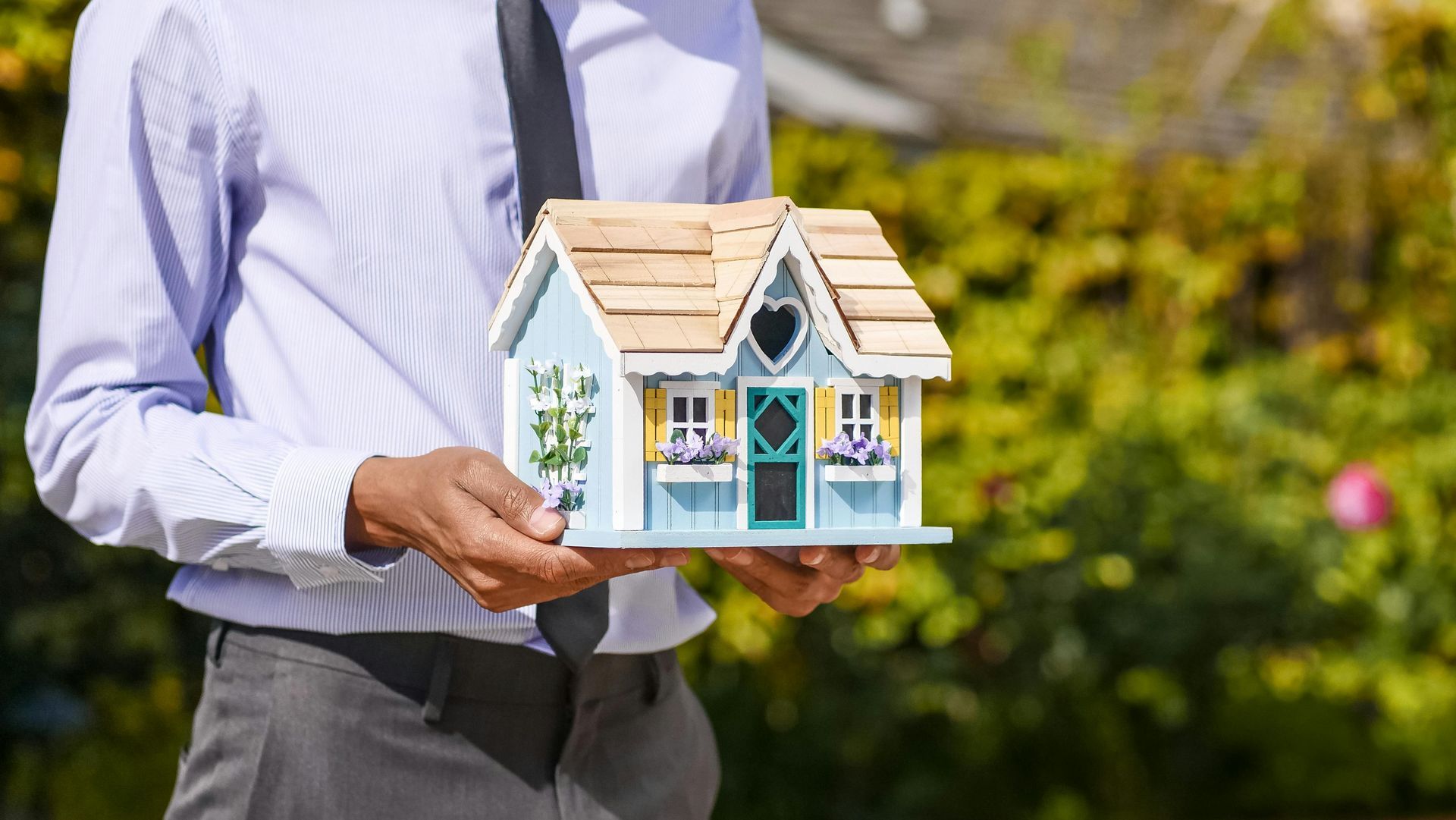 Person holding a small model house outdoors in a sunlit garden