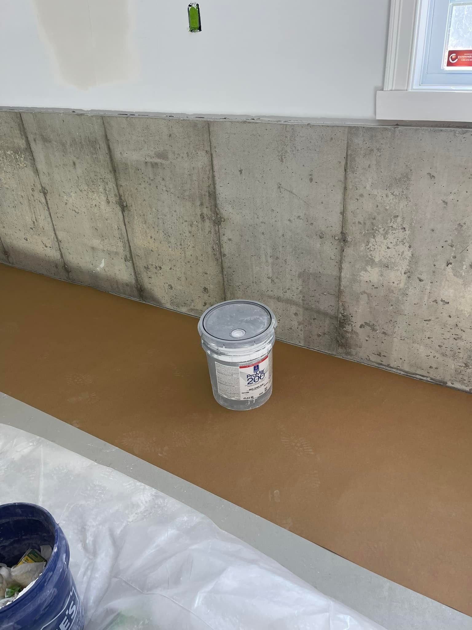 Concrete wall with a paint bucket on a brown surface, with a blue bucket in the foreground.