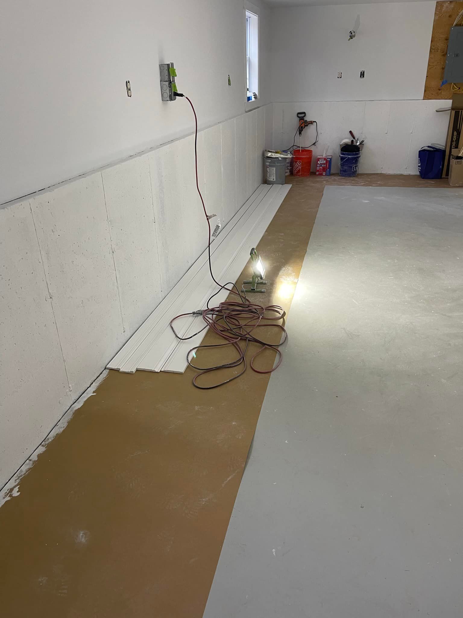 Interior of a room under construction. White walls, gray and brown floors, exposed wires and tools.