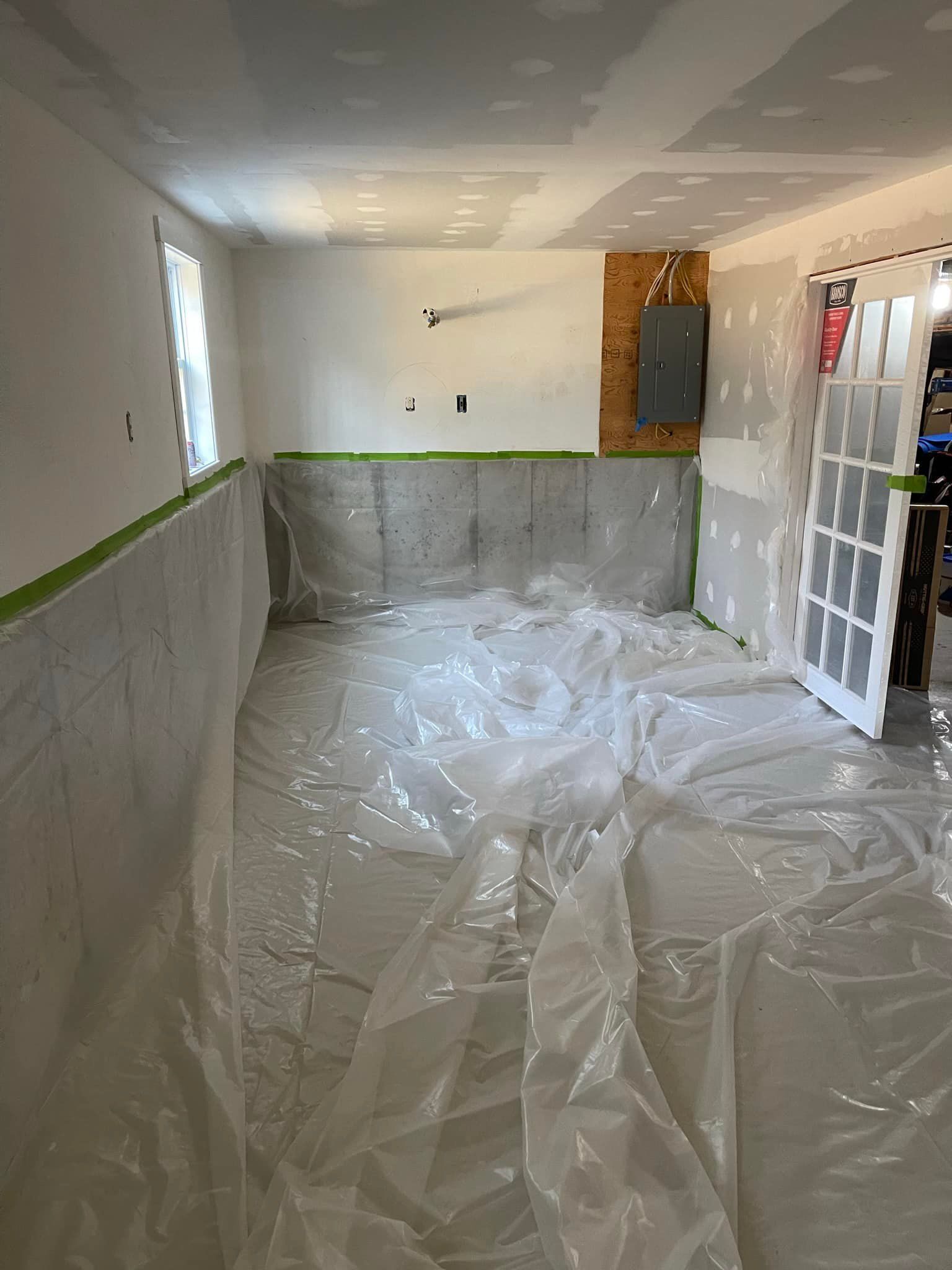 Basement with plastic sheeting on the floor and unfinished walls; door and electrical panel visible.