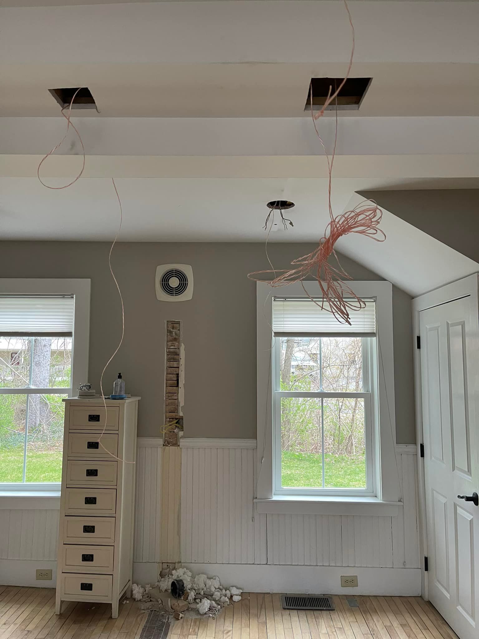 Room under construction, exposed wires hang from ceiling, windows, dresser, door, rubble on floor.