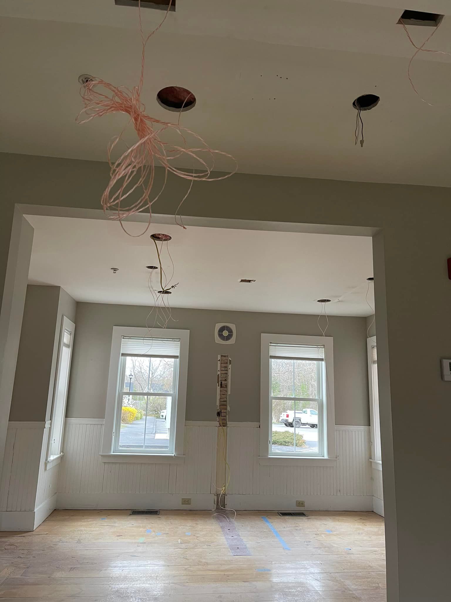 Interior view of a room under construction. Exposed wires hang from the ceiling; windows, trim, and unfinished floors are visible.