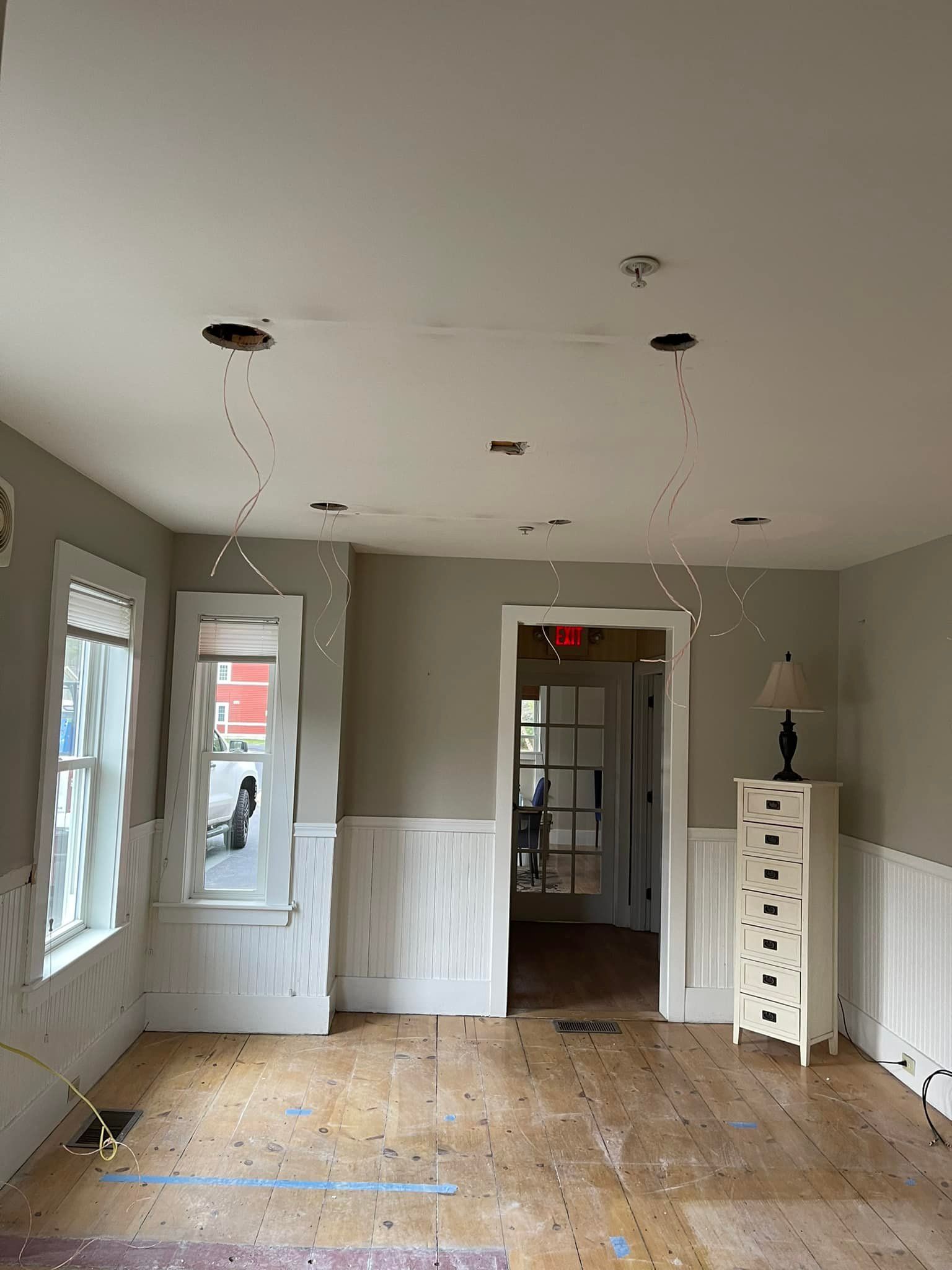 Empty room with light fixtures. White paneling on lower walls, wood floor. Two windows on the left.