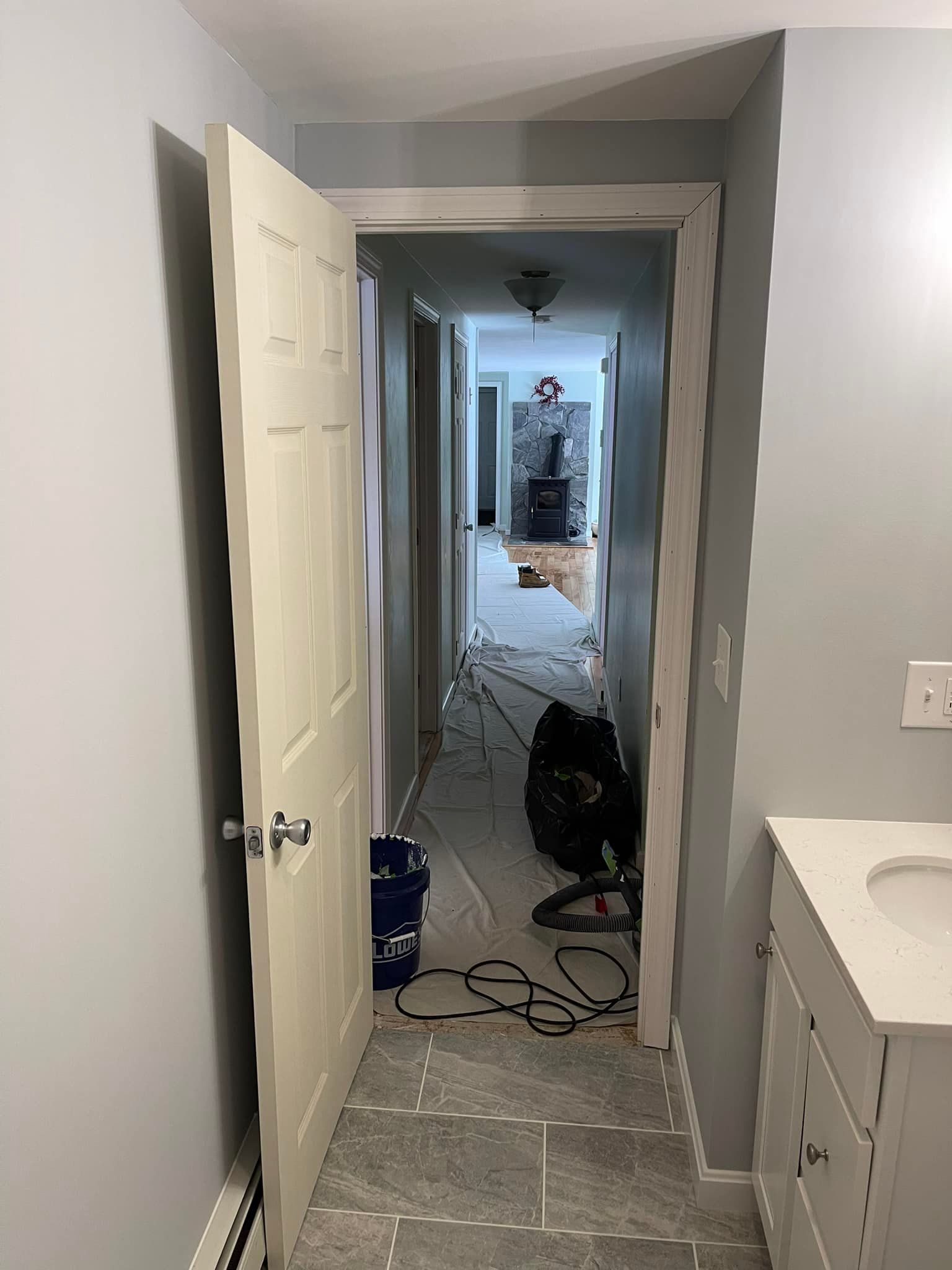 A narrow hallway under construction. Open white door on the left. Grey walls and floor with tools and debris.