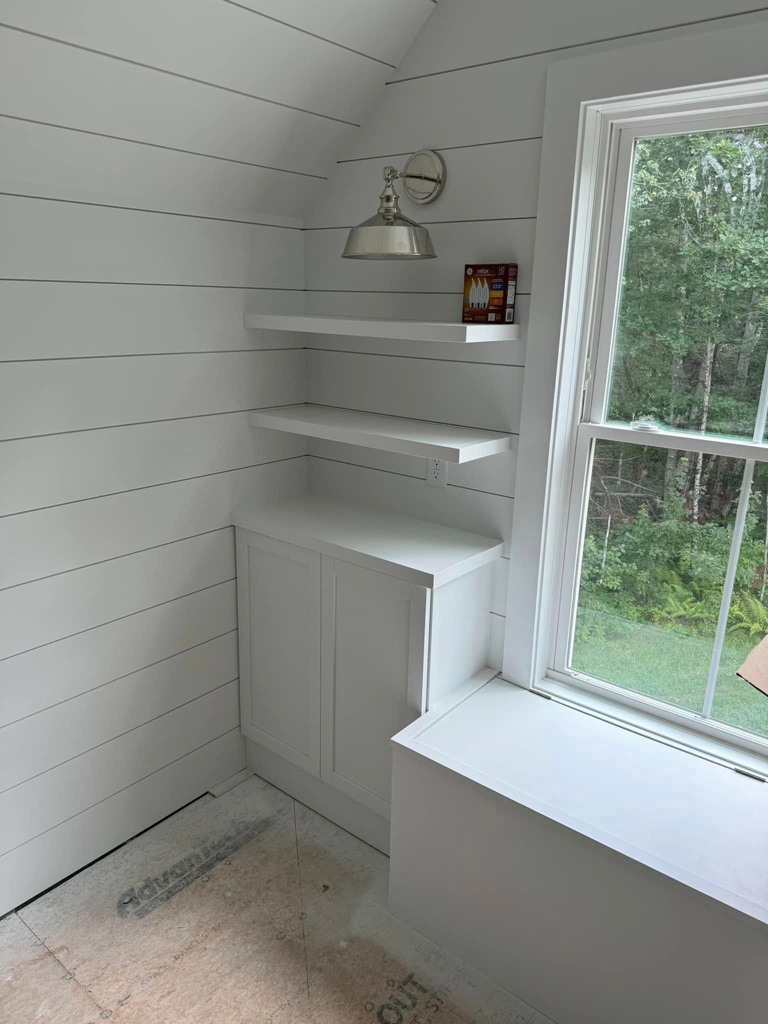 White room corner with shelves, cabinet, window, and wall lamp.