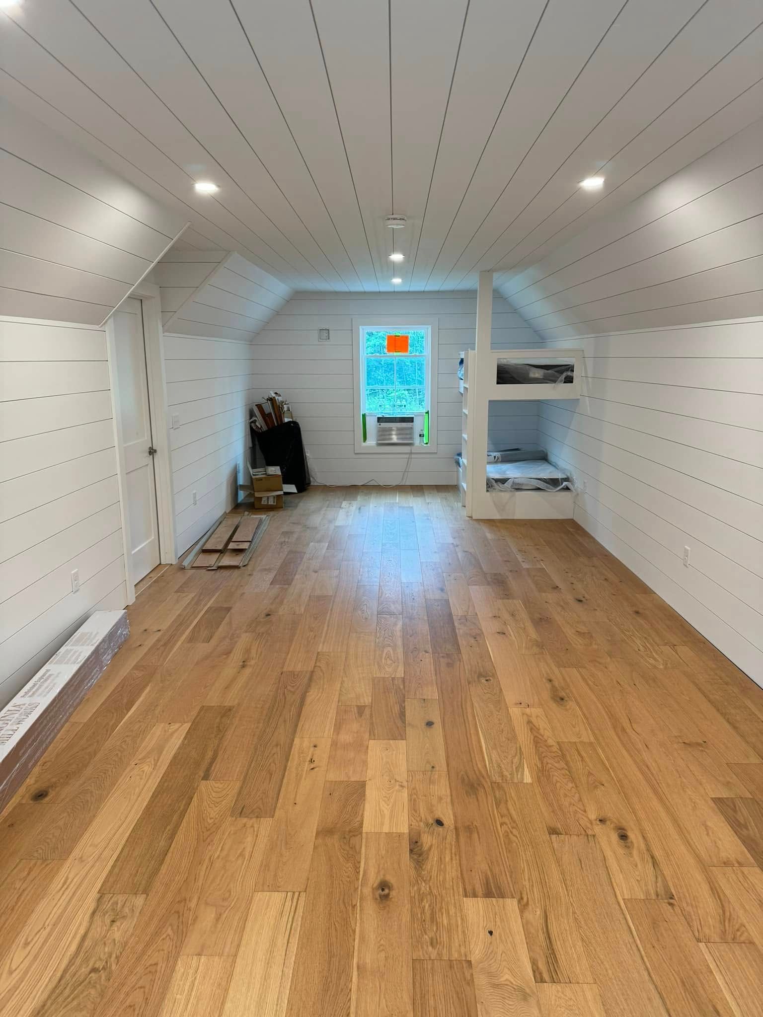 A bright, empty room with light wood floors, white walls, and a window with a bunk bed area.