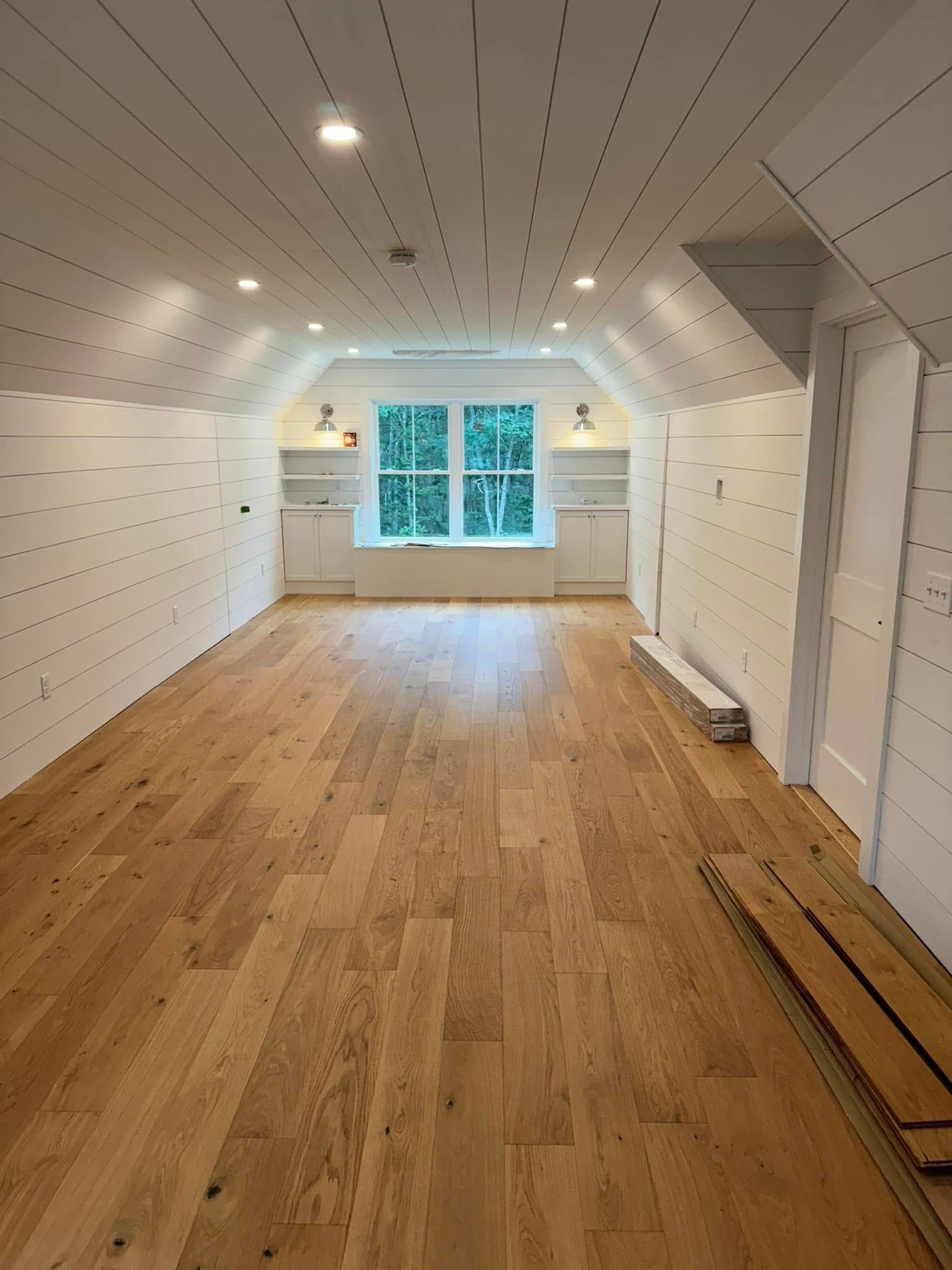 Empty room with wood floors, white paneled walls and ceiling, centered window, and recessed lighting.
