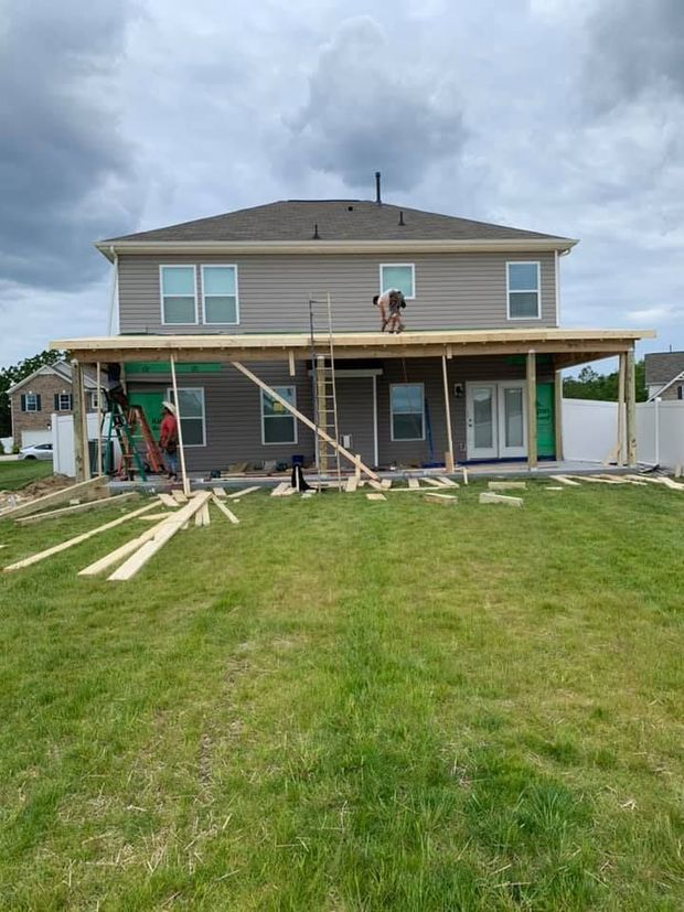Backyard construction: workers building a covered patio on a two-story house. Green grass, cloudy sky.