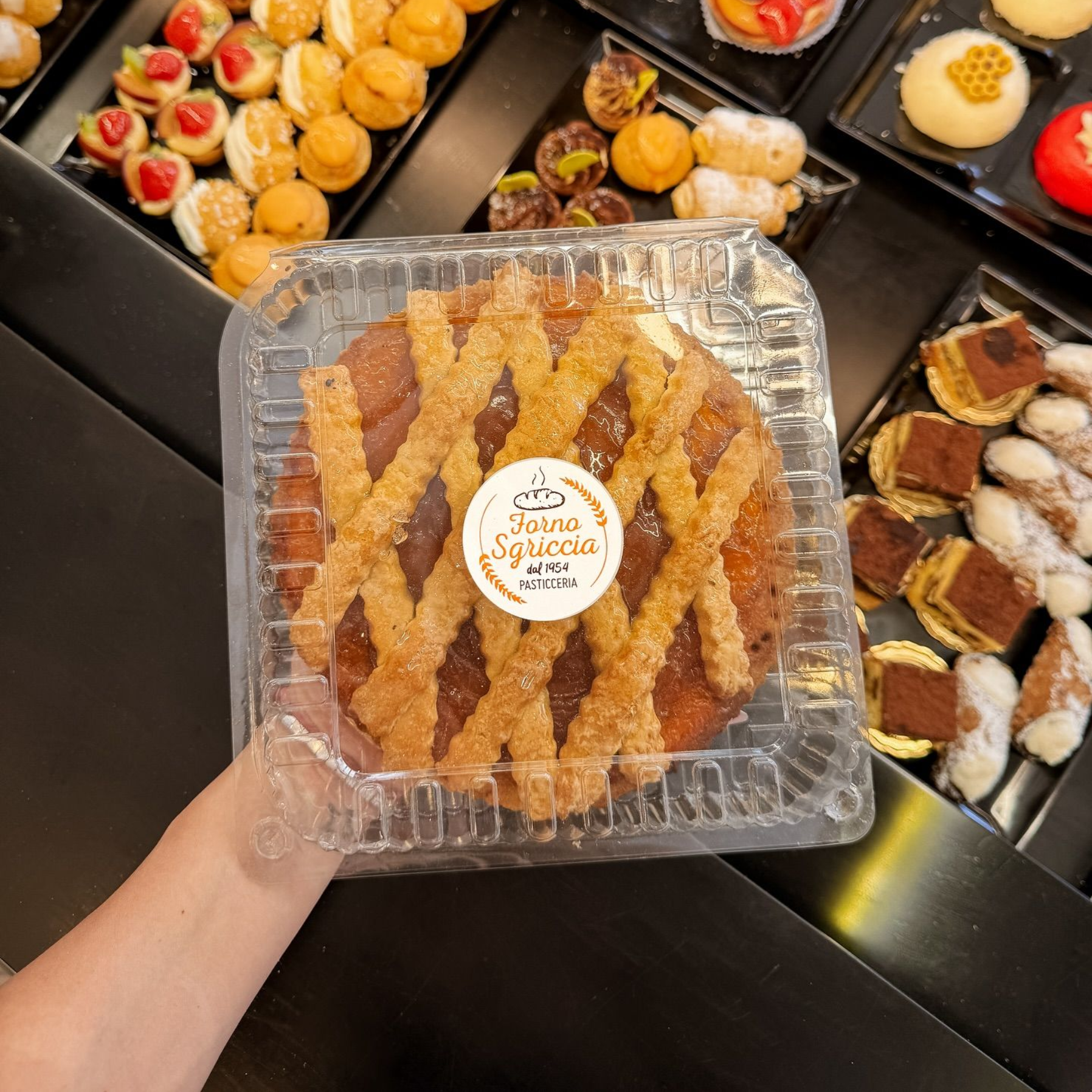 Una donna dietro un bancone regge un piatto di pasticcini. In primo piano, una pila di pasticcini dorati e rettangolari.