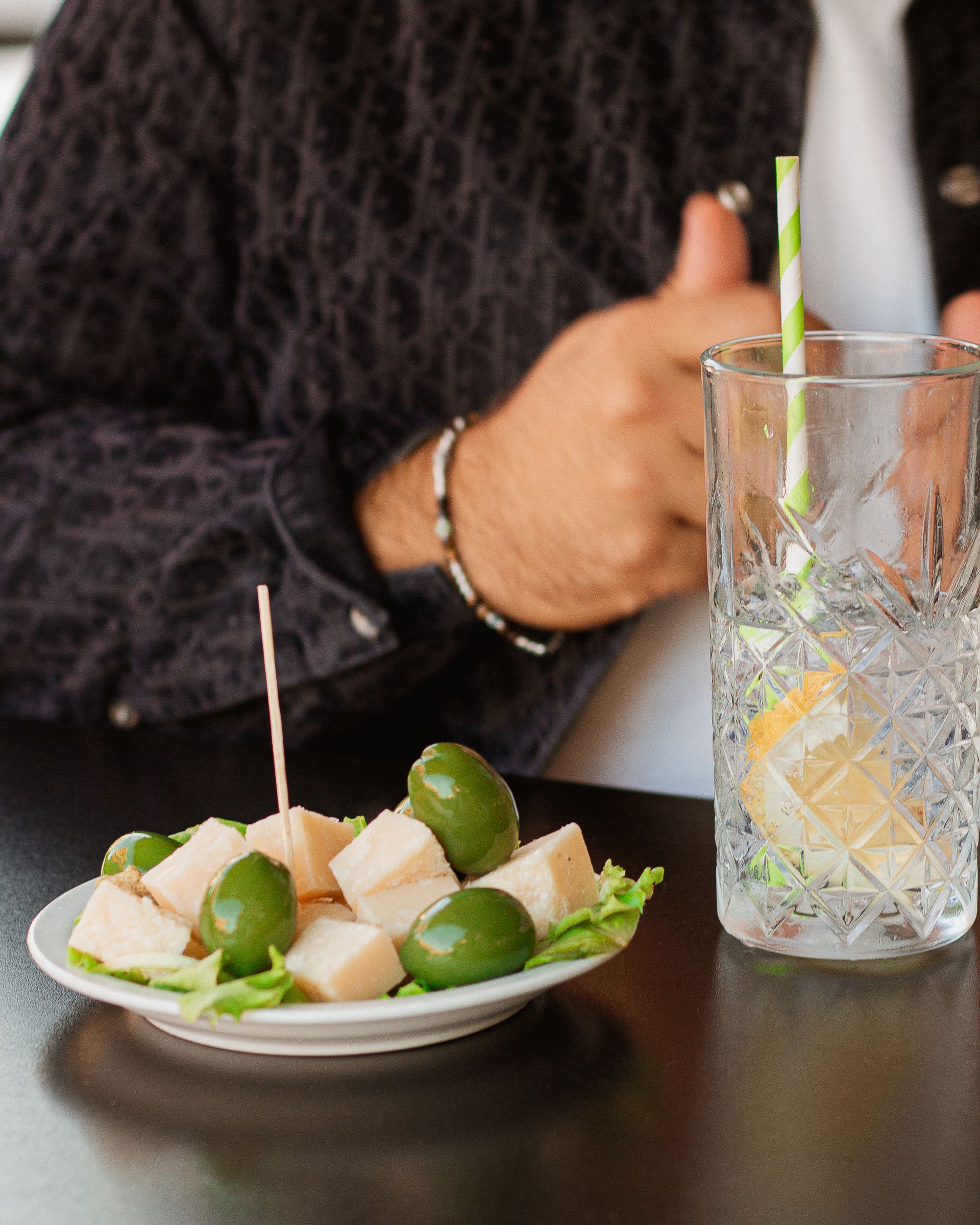 Persona con bevanda e spuntino: un piatto di olive e cubetti di formaggio, un bicchiere con limone, una mano con il pollice alzato.