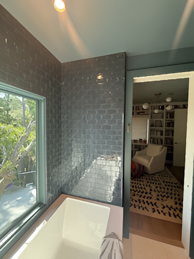 Bathroom with tiled wall, tub, and window overlooking trees. Doorway leads to a room with bookshelf and chair.