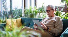 An elderly man is sitting on a couch using a tablet computer.