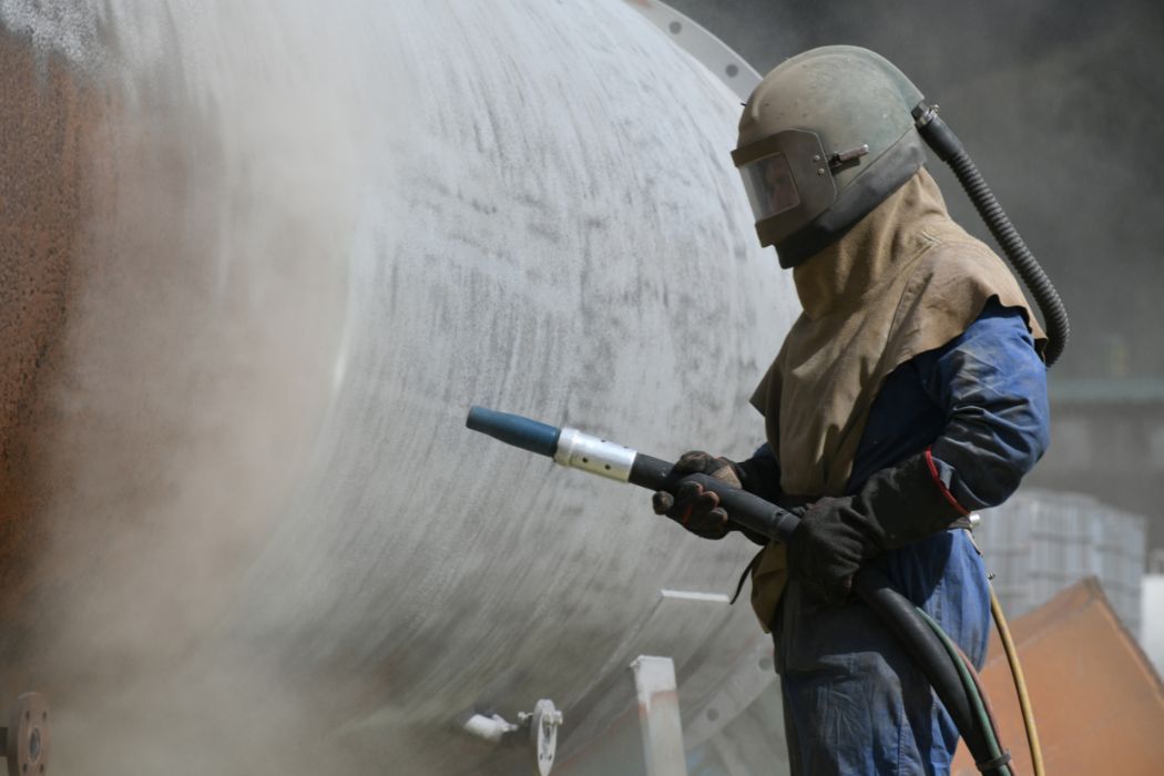 A worker in protective gear and helmet uses a sandblasting nozzle to clean a large industrial metal tank.
