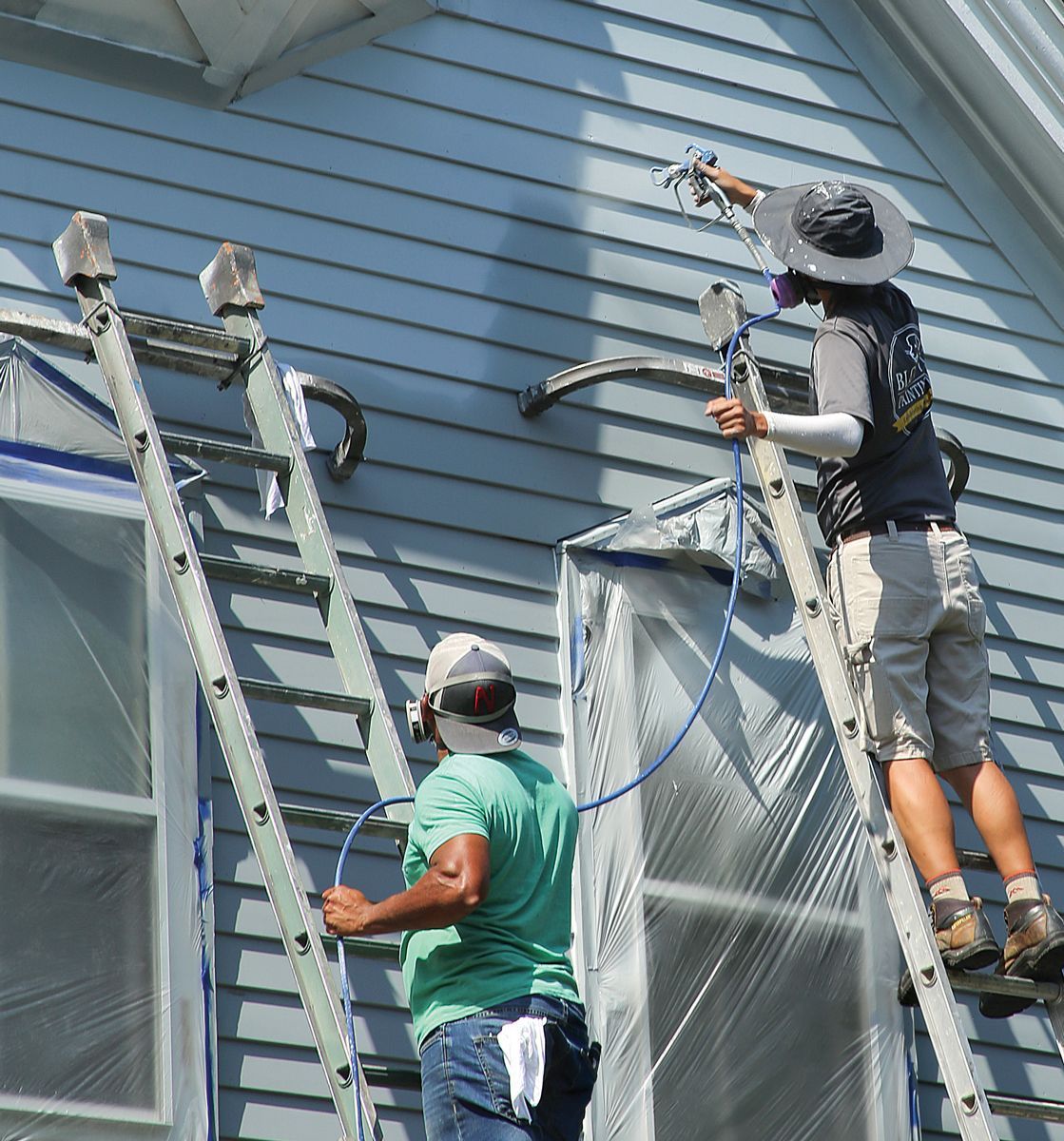Two workers on ladders use paint sprayers to apply a light blue coat to the side of a house.