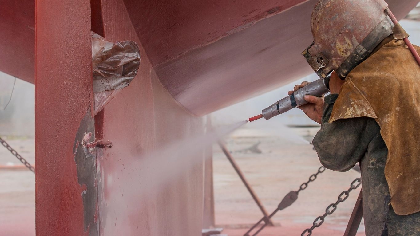 A person in protective gear uses a high-pressure sandblaster to clean a large red metal structure.