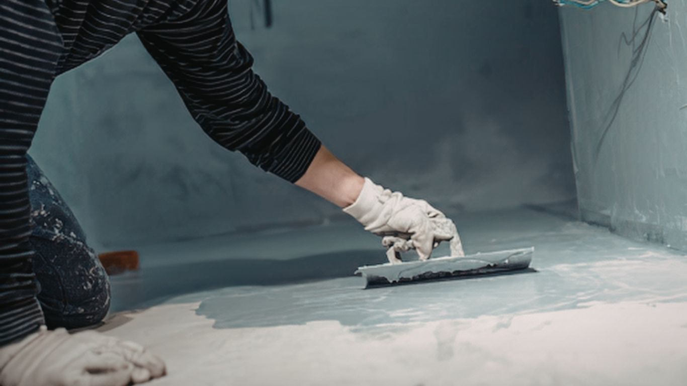 A person wearing gloves uses a trowel to apply light blue mortar or coating to a floor in a construction setting.