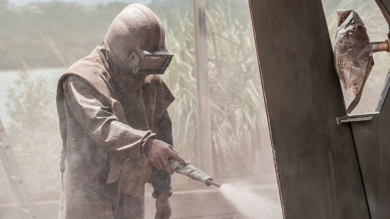 A person in protective gear and a helmet uses a sandblasting tool to clean a metal structure in an outdoor setting.
