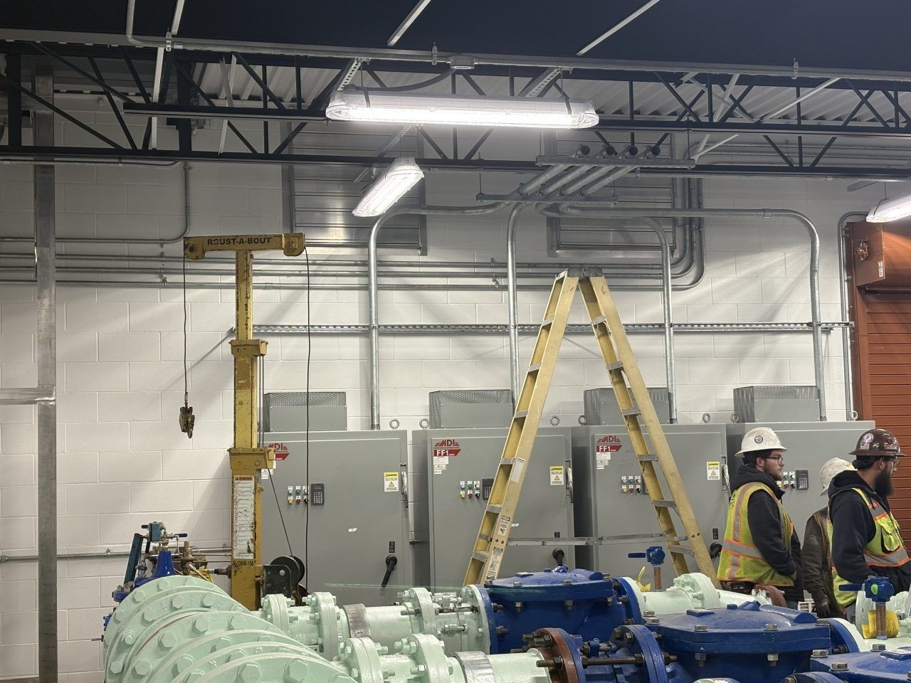 Construction workers in hard hats and vests work near large electrical panels and blue industrial machinery in a warehouse.
