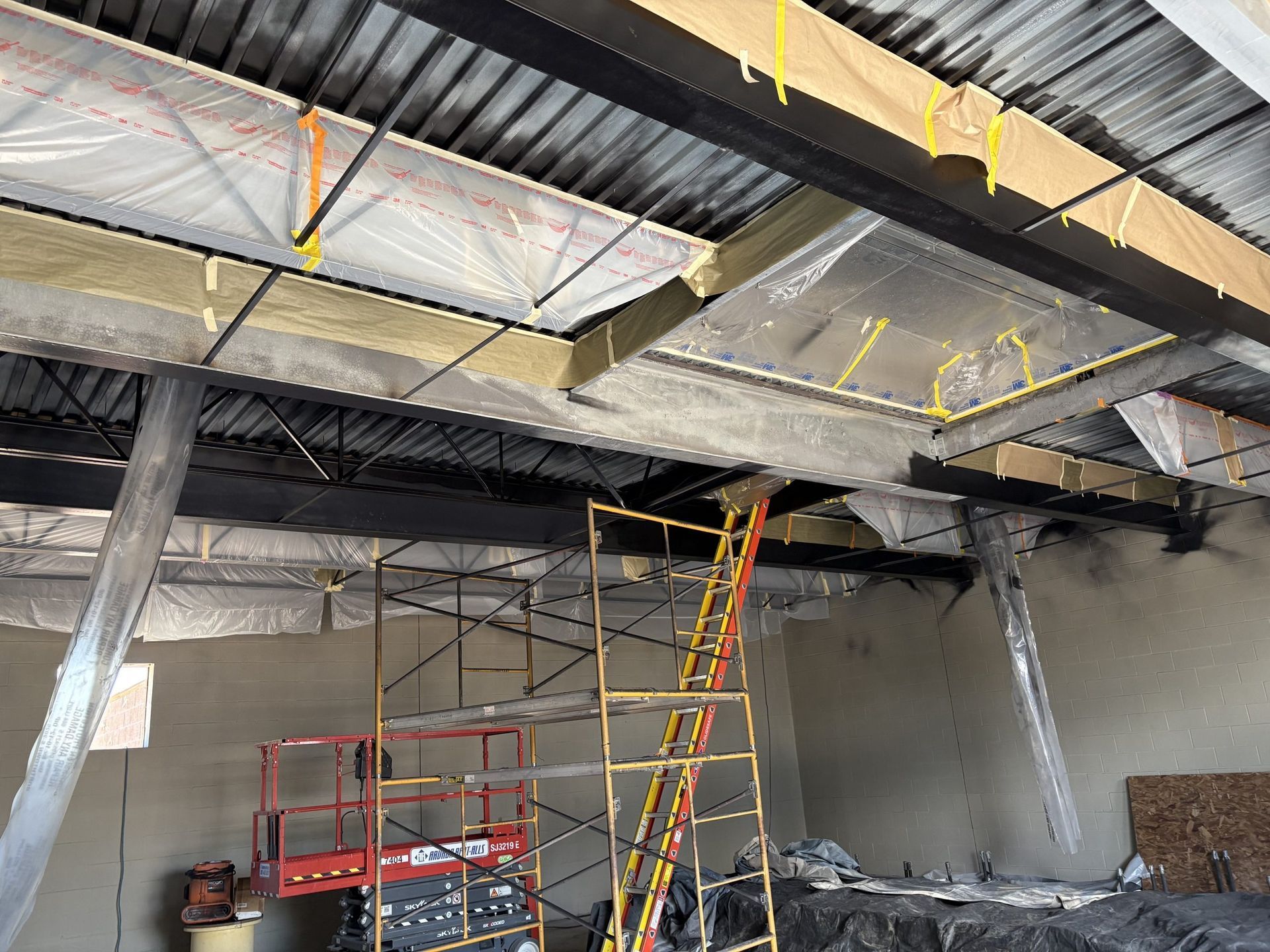 A construction site interior featuring steel beams, metal ceiling decking, insulation, scaffolding, and a scissor lift.