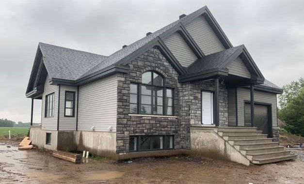 Une grande maison avec une façade en pierre est en construction par une journée nuageuse.