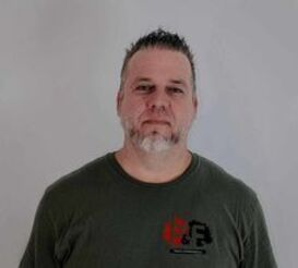 Un homme avec une barbe portant un t-shirt vert se tient devant un mur blanc.