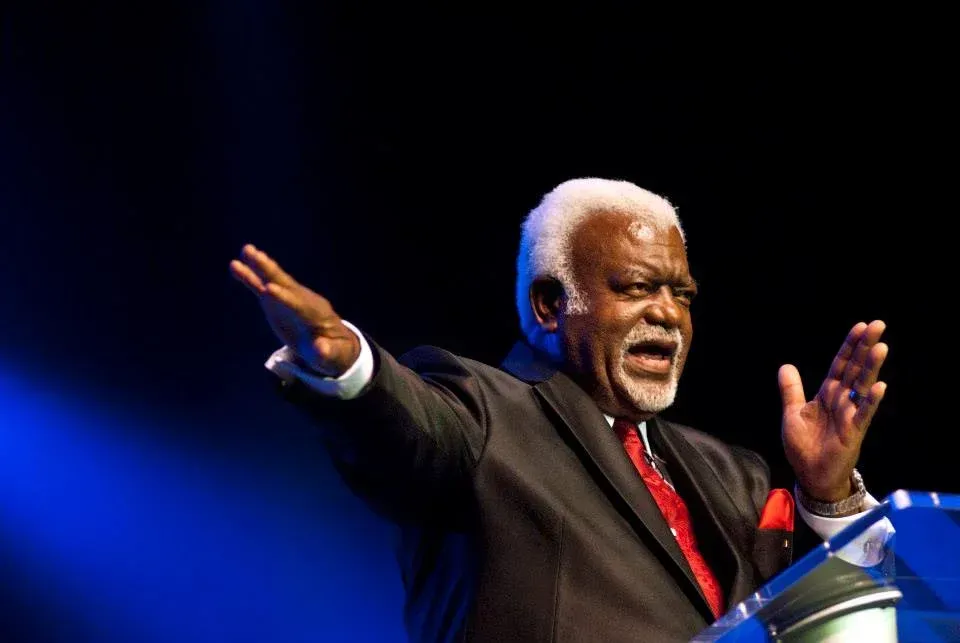 A man in a suit and tie is standing at a podium with his arms outstretched.