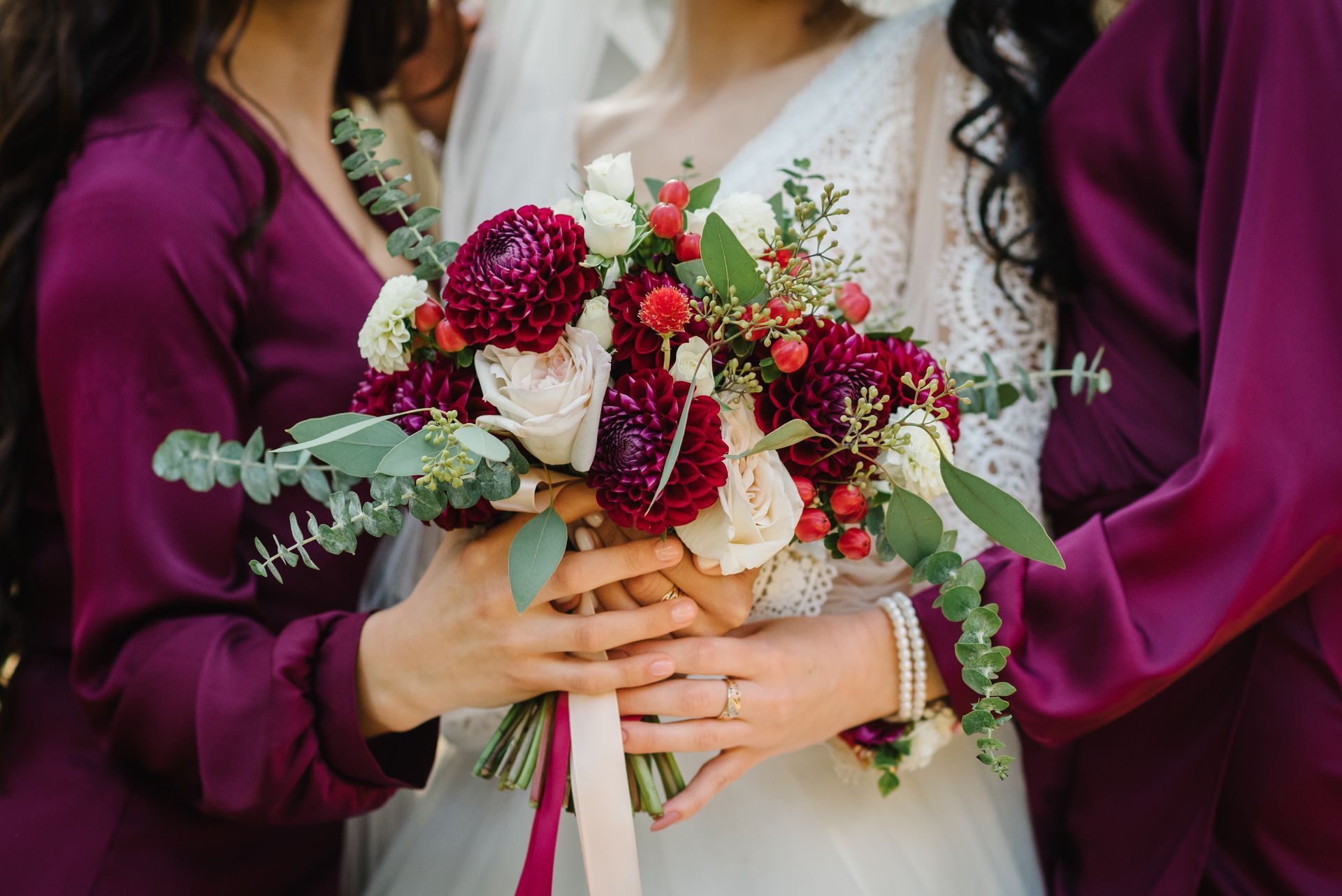 Bride and her bridesmaids' hands holding a bouquet tied with burgundy ribbon