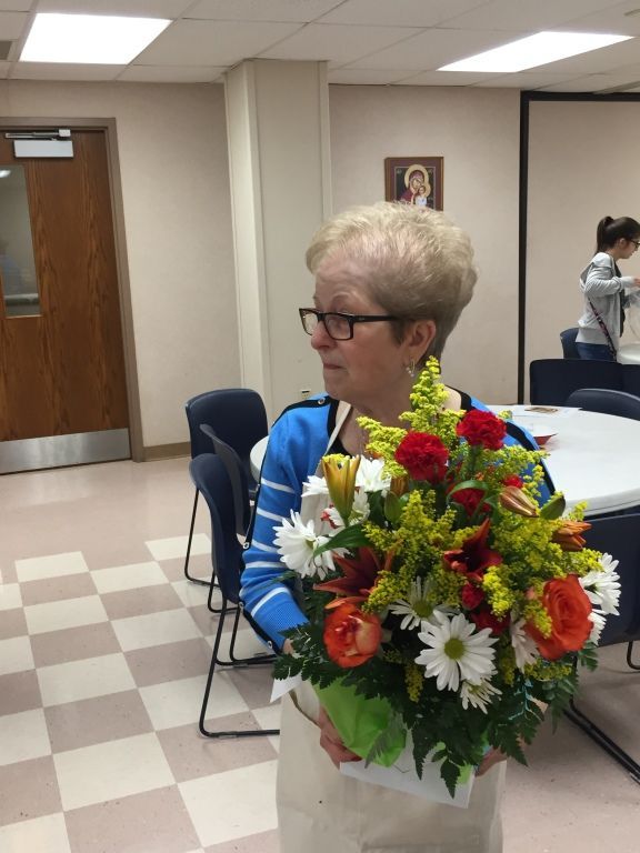 An elderly woman with glasses holds a colorful floral bouquet.