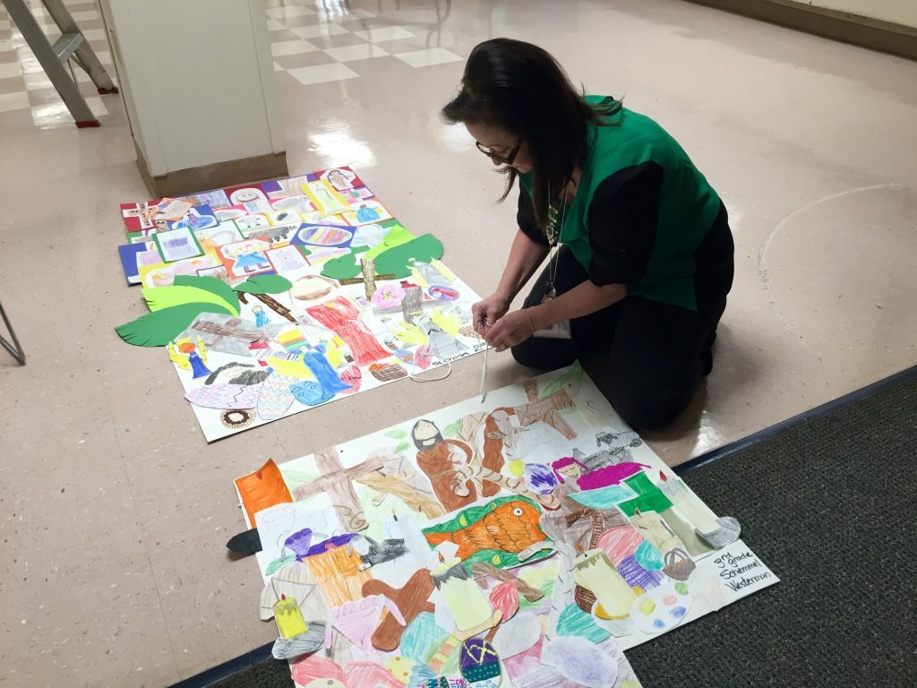 A woman kneels on a tiled floor, arranging colorful, childlike drawings. 