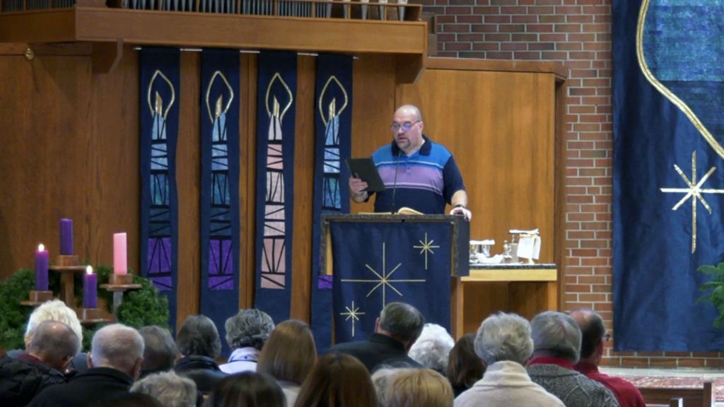 A man stands at a lectern in a church, decorated with blue banners featuring candle designs.