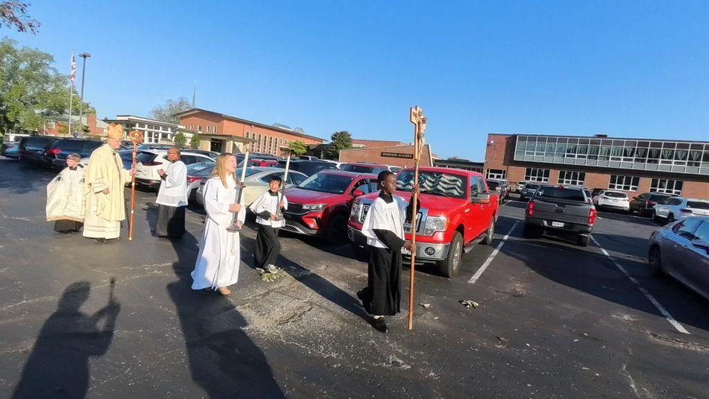 A group of people in religious robes walk through a parking lot, holding a large cross and other ceremonial items.