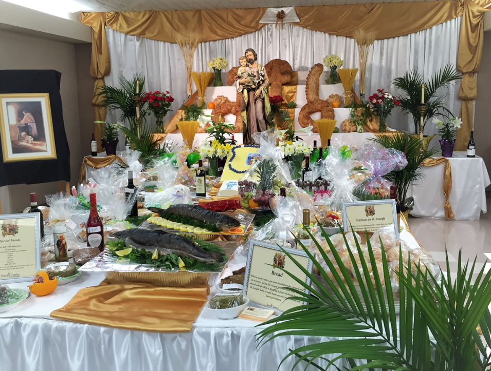An altar adorned with a statue of a saint is surrounded by an array of food and drink offerings. 