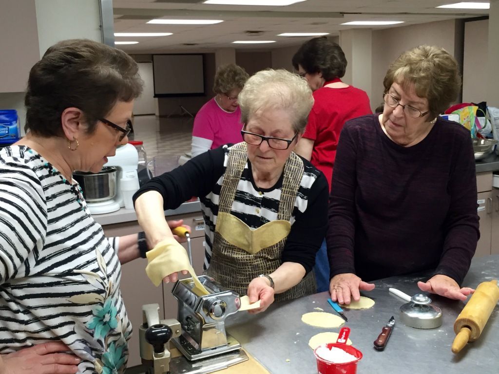 Three elderly women in a kitchen make pasta together, using a hand-crank machine. 