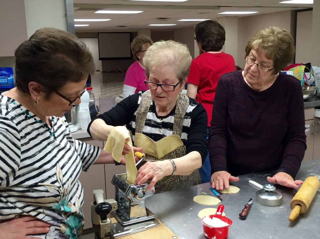Three women collaborate enthusiastically on pasta-making in a kitchen.