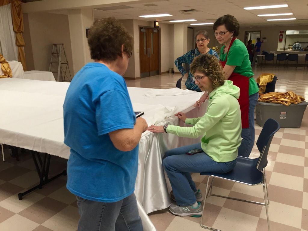 Four women prepare a table covered with a white cloth in a spacious hall. 