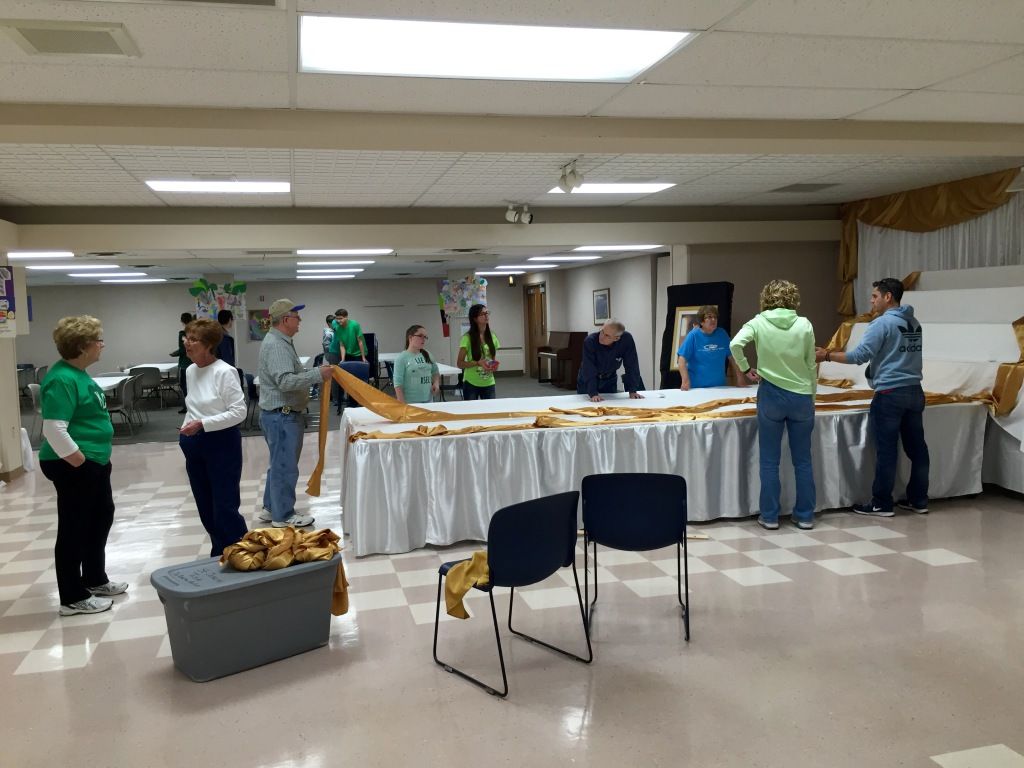 A group of people are setting up gold and white decorations in a large room with tile flooring.