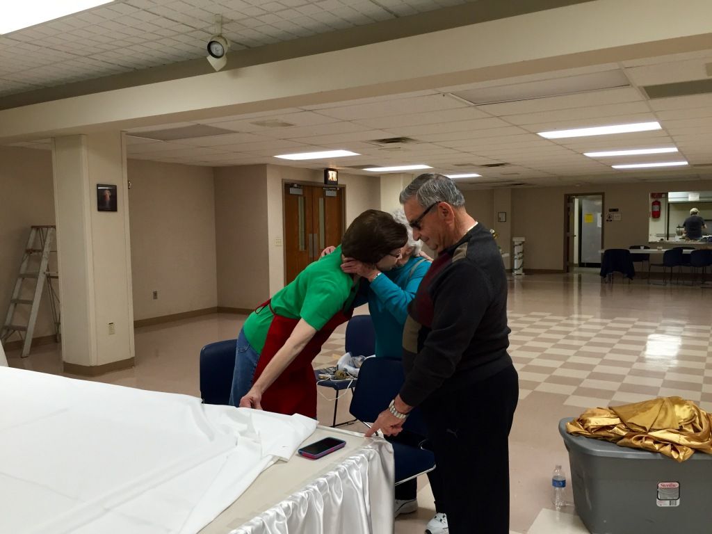 Three people in an empty hall, focused on arranging a white tablecloth on a long table.