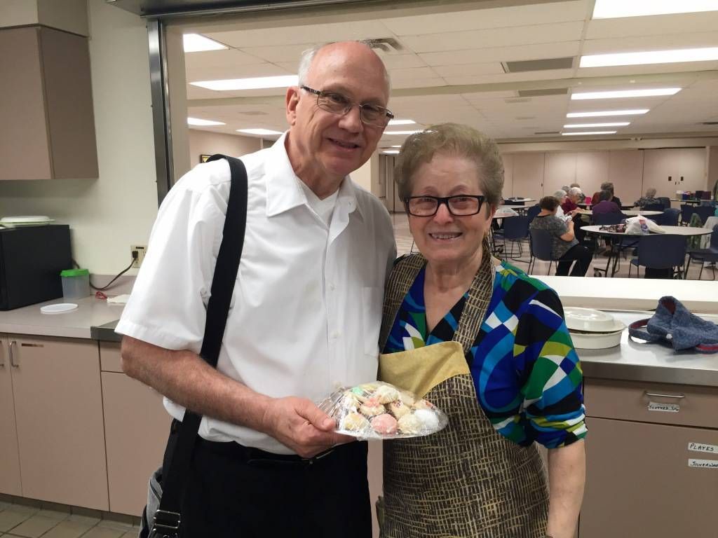 A smiling man and woman stand in a kitchen, holding a plate of colorful cookies. 