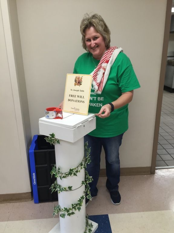A smiling person in a green shirt stands next to a white donation box.