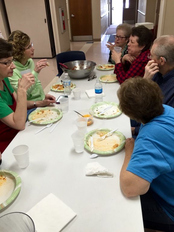 A group of six people sit around a white table enjoying a meal, chatting and smiling. 