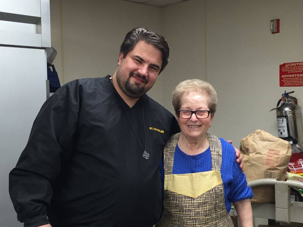 A smiling man in a black jacket stands beside an older woman in a blue and yellow apron.