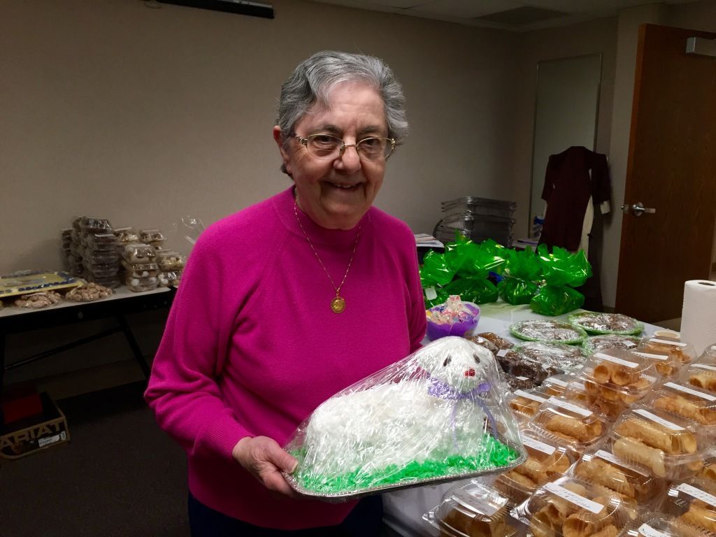 Elderly woman in a pink sweater smiles, holding a lamb-shaped cake wrapped in plastic. 