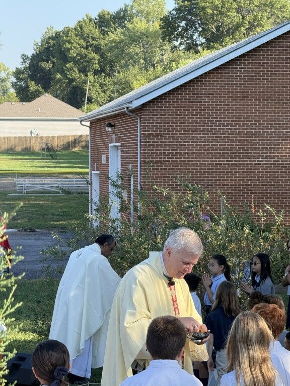 A priest in a yellow robe gives communion to children outdoors near a brick building. 