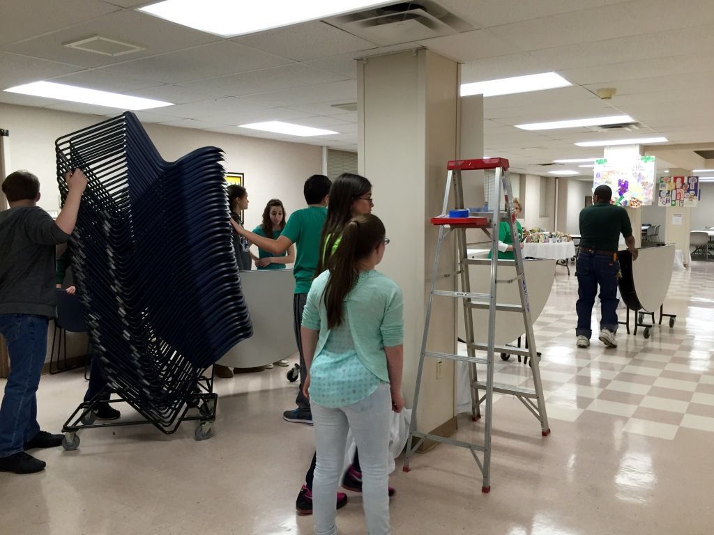 A group of people, work together in a hall setting up chairs and tables.