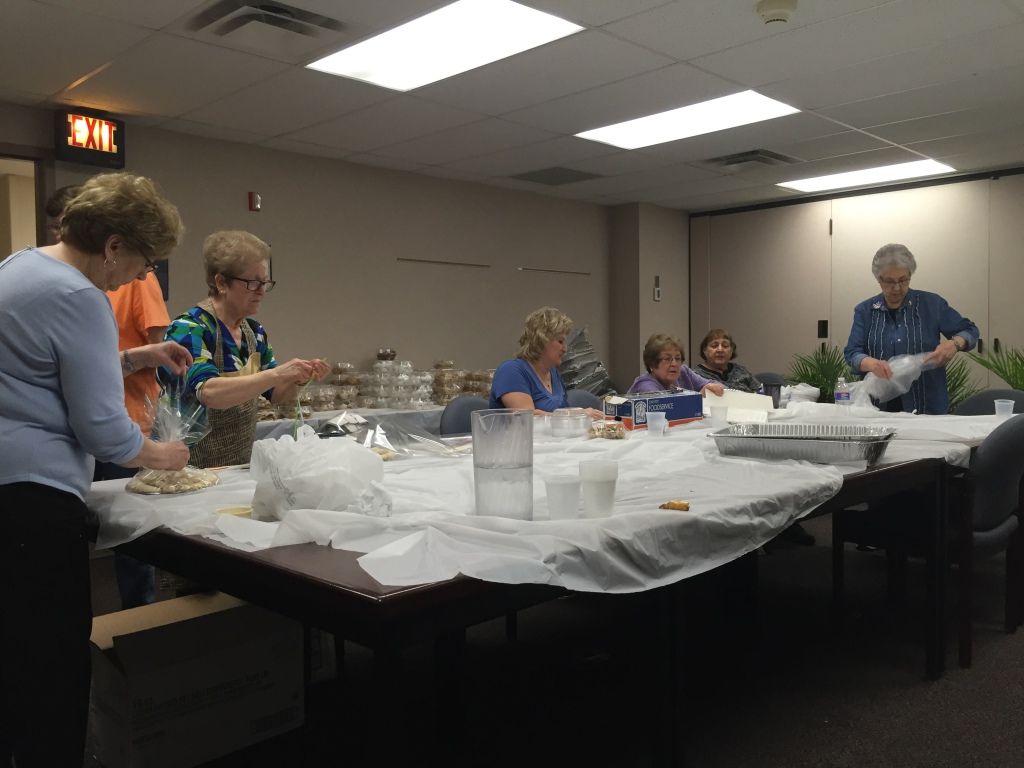 A group of women in a meeting room are gathered around a table covered in a white cloth, packaging baked goods. 