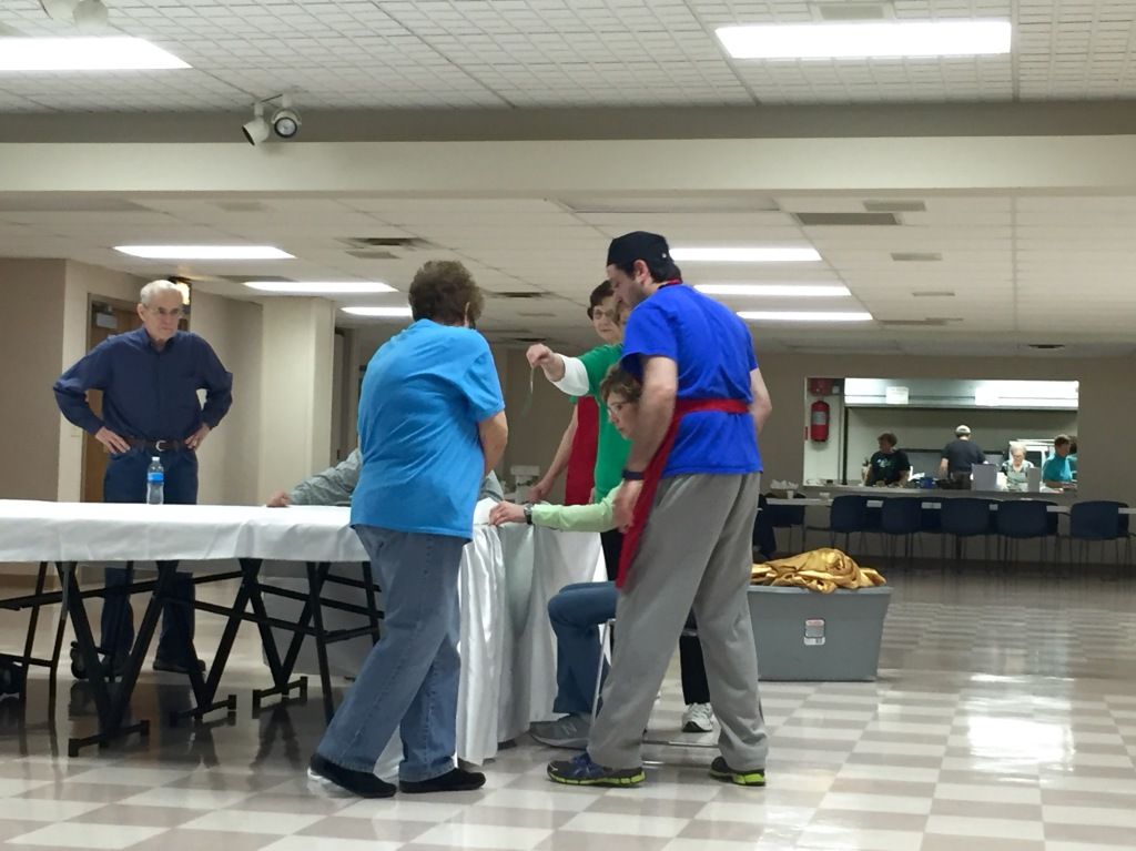 A group of people in a large room prepare a table with white cloths.