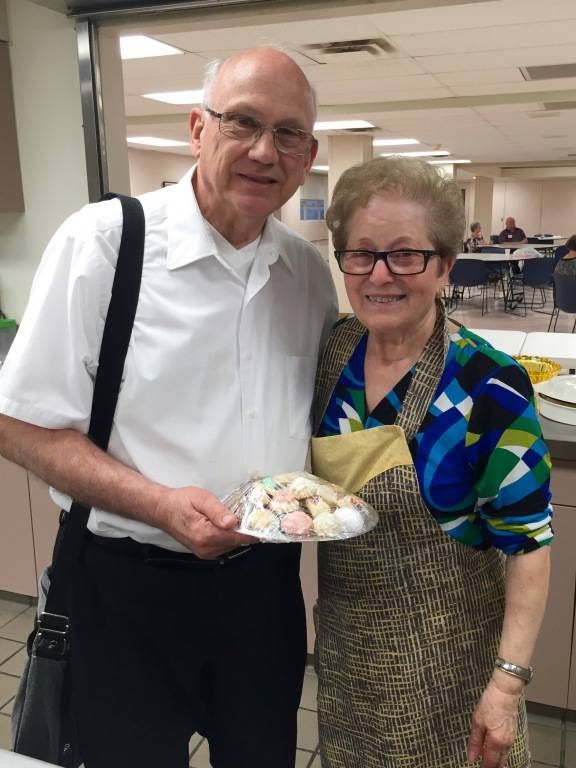 A smiling elderly man and woman stand in a dining hall. 