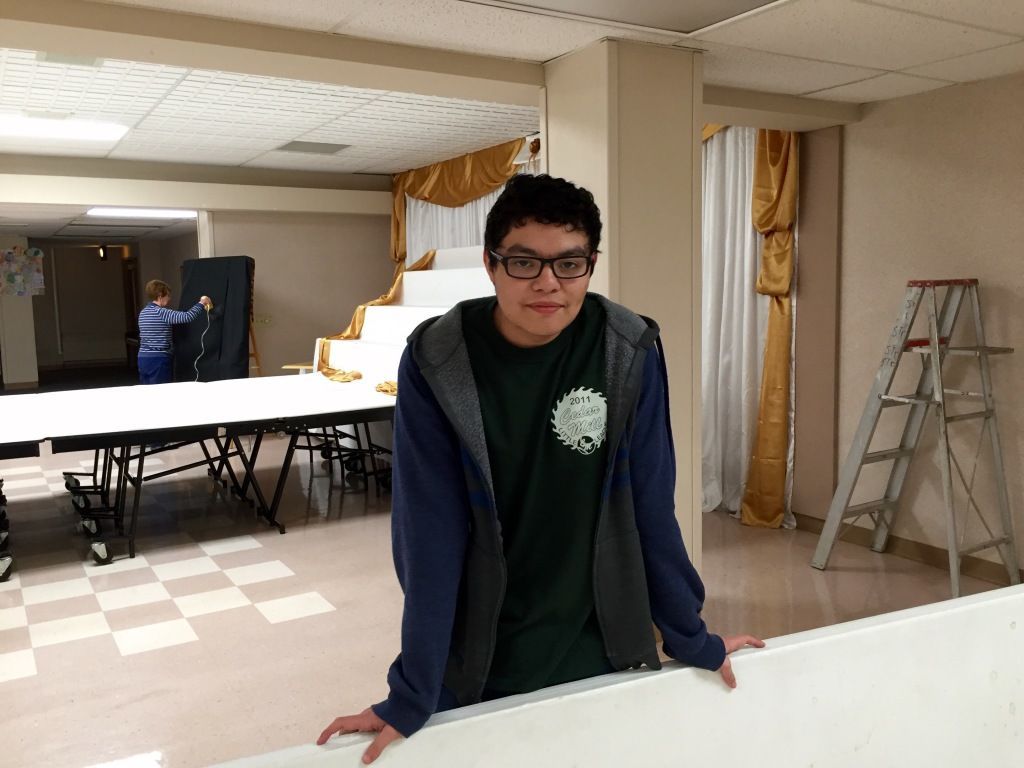 Young person in a blue hoodie leans against a table in a room with gold curtains. 