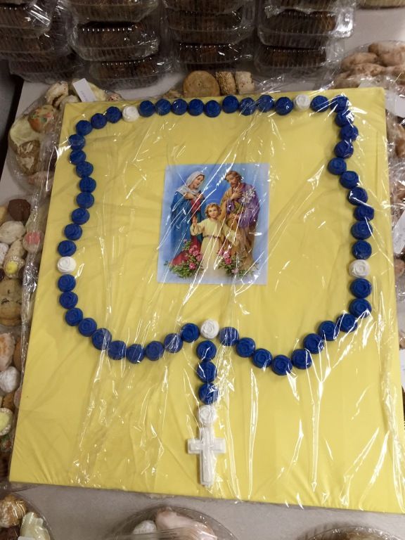 A decorative cake with a yellow surface, featuring a blue and white rosary design and a small image of the Holy Family at the center.
