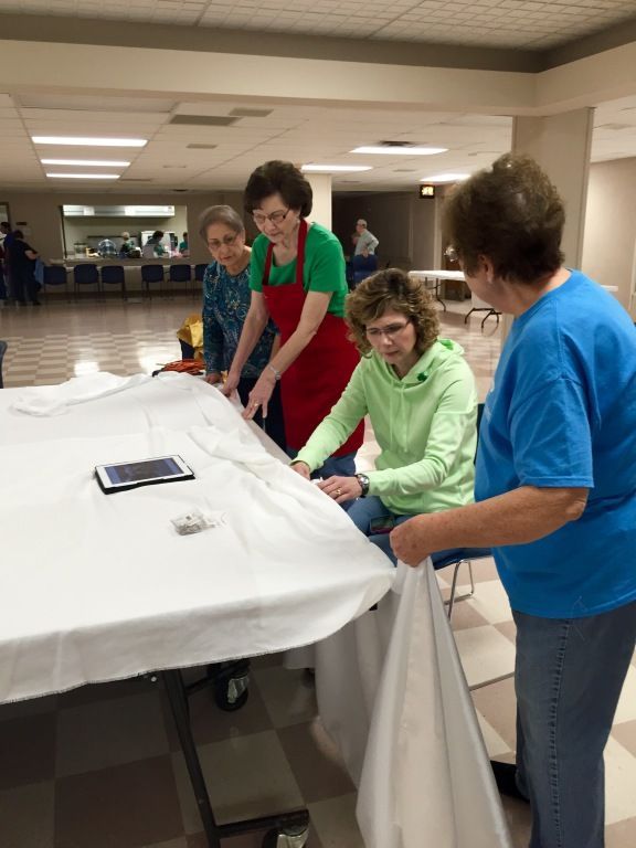 Four women collaborate on sewing, gathered around a table with white fabric in a spacious room.